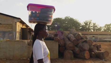 Eine-Junge-Frau-Mit-Einer-Last-Auf-Dem-Kopf-Geht-Nach-Rechts-An-Einem-Sägewerksgelände-Entlang-Und-Verkauft-Möglicherweise-Ein-Paar-Getränke-An-Die-Gemeinde-In-Kumasi,-Ghana