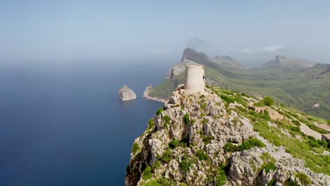Drone-flight-past-an-old-ruined-tower-on-the-top-of-a-mountain-in-a-hilly-island-landscape-surrounded-by-blue-ocean