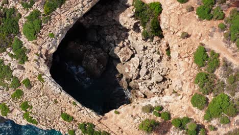 Birds-eye-view-on-cave-on-the-edge-of-a-cliff-with-strong-waves-breaking-on-the-shore,-Mediterranean-landscape,-aerial-view