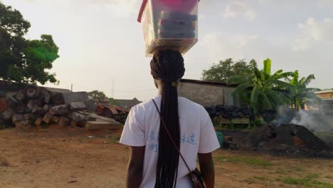 Young-woman-seen-from-the-back-revealing-her-long-beautiful-hair,-a-plastic-box-on-her-head,-walking-through-a-sawmill-compound,-must-be-peddling-drinks-to-the-workers-and-the-village,-Kumasi,-Ghana