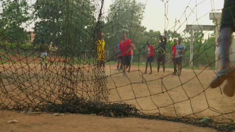 Torwart,-Gesehen-Durch-Das-Netz,-Wie-Er-Nach-Rechts-Springt-Und-Den-Ball-Fängt,-Während-Junge-Fußballspieler-Abwechselnd-Freistöße-In-Richtung-Tor-üben,-Kumasi,-Ghana