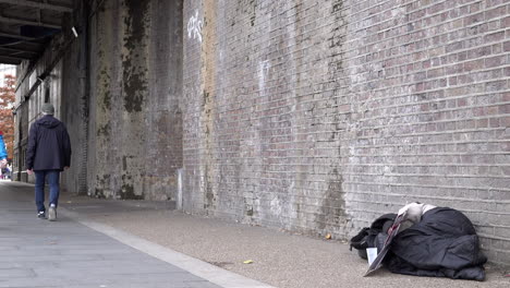 People-and-pigeons-walk-past-a-homeless-person-asleep-in-a-grey-sleeping-bag-on-the-floor-of-an-underpass-during-daylight-in-winter