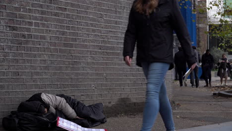A-homeless-person-sleeps-in-a-sleeping-bag-on-the-floor-of-an-underpass,-next-to-a-cardboard-sign-that-reads,-“Hungry-and-homeless”-that-falls-over-as-people-walk-past-during-daylight-in-winter