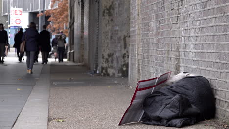 Menschen-Gehen-Bei-Tageslicht-Im-Winter-An-Einem-Obdachlosen-Vorbei,-Der-In-Einem-Grauen-Schlafsack-Auf-Dem-Boden-An-Der-Ziegelwand-Einer-Unterführung-Schläft