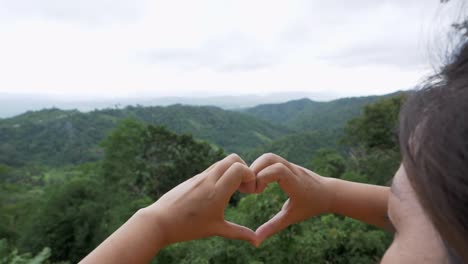 Frau-Hält-Ihre-Finger-Zusammen-Und-Bildet-Eine-Herzform,-Während-Sie-Auf-Die-Berge,-Den-Wald,-Den-Himmel-Und-Alles-über-Die-Natur-Blickt,-Während-Sich-Die-Kamera-Von-Ihrem-Kopf-Zu-Ihren-Händen-Bewegt,-Thailand