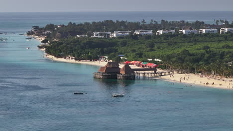 Panorama-of-coastline-at-sunny-day,-drone-view-of-sandy-beach,-turquoise-ocean,-luxury-resort-and-beautiful-thatch-stilt-house-restaurant,-Zanzibar,Tanzania