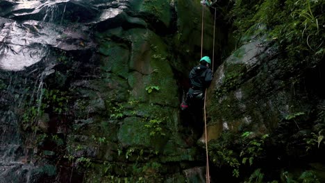 Aerial-view-of-woman-canyoning-on-wet-steep-mountain-wall-beside-waterfall
