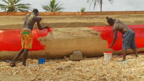 couple-of-workers-working-as-a-team-painting-a-red-wooden-fisherman-boat-on-a-tropical-sand-beach-in-Africa