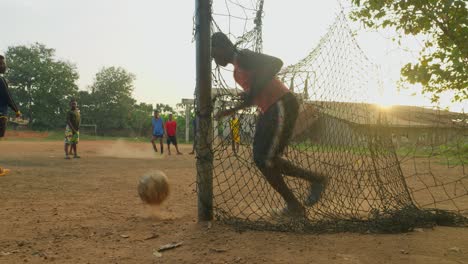 Precioso-Escenario-De-Hombres-Jóvenes-Jugando-Al-Fútbol-Mientras-El-Portero-Impide-Que-El-Balón-Entre-En-La-Portería-Mientras-Las-Puestas-De-Sol-Crean-Una-Silueta-En-Un-Campo-De-Fútbol-Comunitario-En-Kumasi,-Ghana.