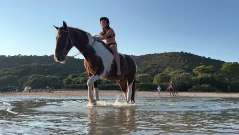 Low-angle-view-of-little-girl-with-equestrian-cap-riding-horse-in-sea-water-on-the-beach-in-summer-season