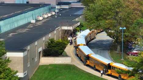School-buses-lined-up-outside-a-building-with-students-and-teachers-walking-nearby,-surrounded-by-greenery