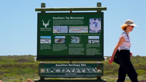 Tourists-walk-past-Southern-Tip-Monument-information-board,-Cape-Agulhas-tourism