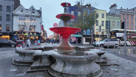 Straße-In-Cork-City-Mit-Rot-Beleuchtetem-Wasserbrunnen,-Fußgängern,-öffentlichen-Verkehrsmitteln,-Verkehr,-Abendzeit-Im-Winter-Mit-Weihnachtsdekorationen