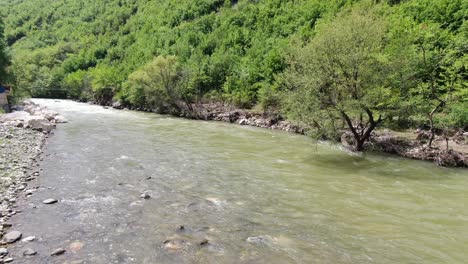 Drone-view-in-Albania-flying-over-a-moving-river-between-green-mountains-on-a-sunny-day