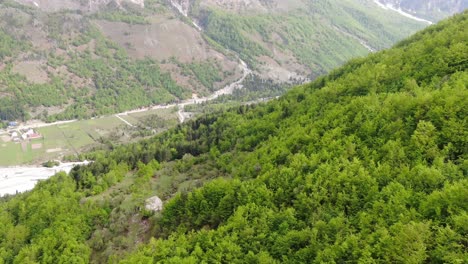 Drone-view-in-Albania-flying-in-the-alps-showing-green-forest-on-a-valley-surrounded-by-mountain-with-snowy-peaks-in-Valbon?