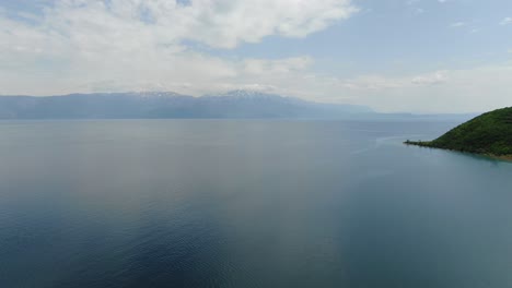Drone-view-in-Albania-flying-over-Ohrid-blue-lake-with-snowy-mountains-in-the-horizon-and-green-forest-on-the-side