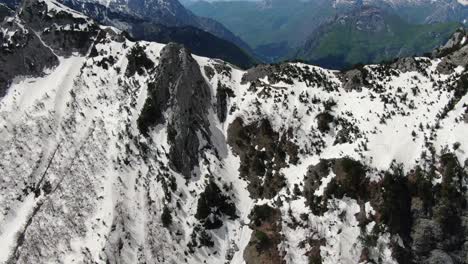 Drone-view-in-Albania-in-the-alps-flying-over-a-snowy-and-rocky-mountain-peak-and-into-a-green-forest-hill-on-the-side-in-Theth