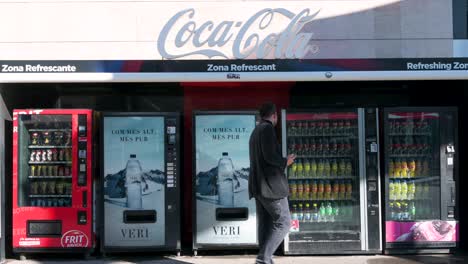 Un-Comprador-Compra-Agua-En-Una-Máquina-Expendedora-De-Bebidas-Mientras-Se-Ven-Numerosos-Bocadillos-Y-Ofertas-De-Refrescos,-Como-Coca-cola-Y-Fanta,-Listos-Para-Su-Compra.