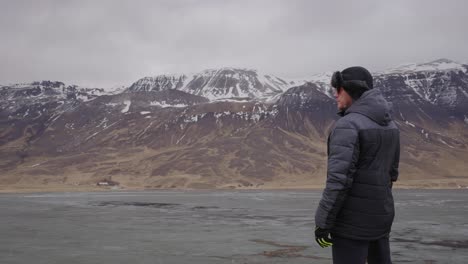 Man-stand-on-cliff-edge-near-lake,-Icelandic-mountain-range-in-background