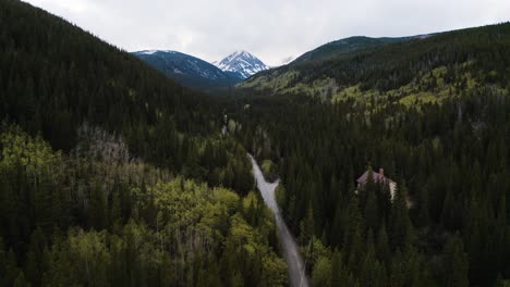 Aerial-shot-tilting-up-to-reveal-a-snow-capped-mountain-nestled-between-green-forests-in-the-Rocky-Mountains