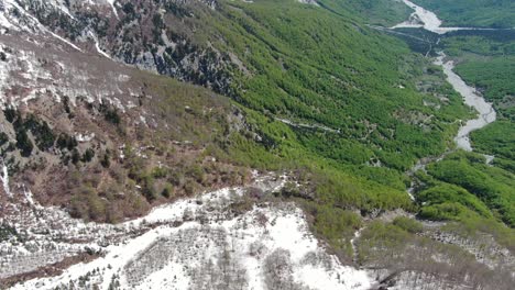 Drone-view-in-Albania-flying-in-the-alps-showing-green-and-snowy-forest-on-a-valley-surrounded-by-mountain-with-snowy-peaks-in-Theth