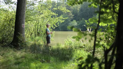 Male-angler-stands-on-shore-of-cloudy-lake-between-green-grass-and-trees-catching-fish-with-fishing-rod,-pan