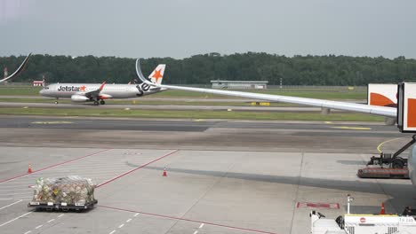 Jetstar-Flugzeug-Rollt-Auf-Der-Landebahn-Im-Hintergrund,-Hinter-Dem-Flügel-Eines-Flugzeugs,-Das-Am-Terminal-Gate-Am-Changi-Flughafen-In-Singapur-Geparkt-Ist