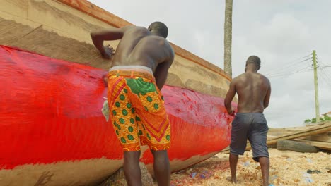 Team-Aus-Zwei-Arbeitern,-Die-Im-Rahmen-Der-Routinemäßigen-Wartung-Ein-Traditionelles-Afrikanisches-Fischerboot-Aus-Holz-Bemalen