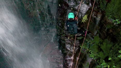 Close-up-shot-of-female-rock-climber-on-rope-beside-waterfall-canyoning-in-rainforest---aerial-view