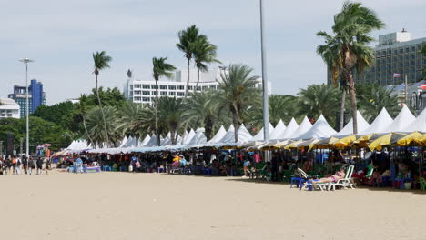 Belebter-Strand-Mit-Menschen-Unter-Vordächern-Und-Sonnenschirmen,-Während-Andere-An-Einem-Windigen-Tag-In-Der-Sonne-Herumlaufen,-Pattaya,-Thailand