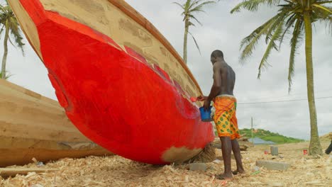 Schwarzer-Männlicher-Afrikanischer-Arbeiter,-Der-Ein-Traditionelles-Hölzernes-Fischerboot-Am-Tropischen-Sandstrand-Von-Ghana-Mit-Palme-Bemalt
