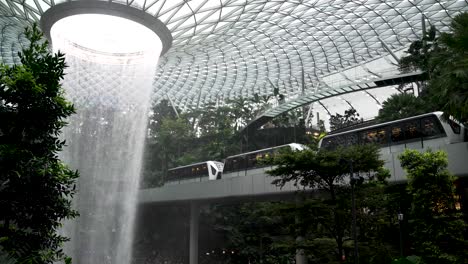 Eine-Zeitlupenaufnahme-Des-Spektakulären-Indoor-Wasserfalls-Namens-„Vortex“-Am-Flughafen-Jewel-Changi,-Im-Hintergrund-Fährt-Der-Sky-Train-Mit-Reisenden-Vorbei,-Singapur