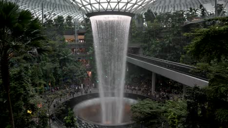 A-slow-motion-shot-of-the-impressive-indoor-waterfall-called-the-Vortex-at-Jewel-Changi-Airport-and-the-surrounding-trees-and-greenery,-Singapore