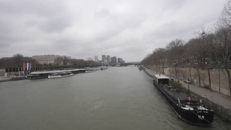 Static-cityscape-shot-of-the-Seine-river-flowing-through-the-city-of-Paris,-boats-anchored-to-the-side,-financial-district-in-the-distance