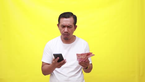 Confused-asian-adult-man-standing-while-holding-cell-phone-and-banknotes