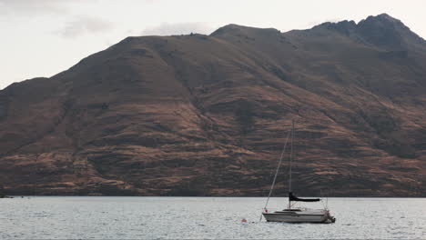 A-sailboat-gently-bobs-in-the-water,-tethered-in-front-of-a-majestic-mountain-backdrop