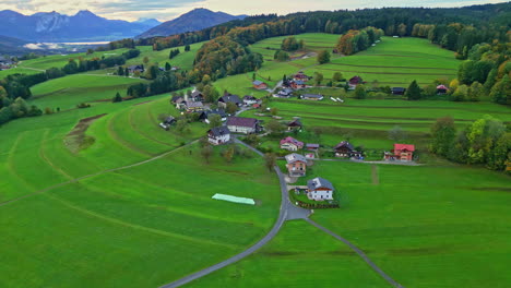 Epic-aerial-view-of-a-scenic-European-village-surrounded-by-lush-green-landscape,-autumn-trees-and-mountains