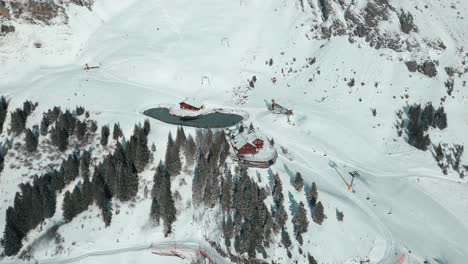 View-from-the-top-of-an-approaching-drone-shot-of-a-cabin-with-a-man-made-lake,-a-cable-stop-by-the-snowy-slope-of-the-Alps-in-Engelberg,-Brunni,-in-Bahnen,-Switzerland