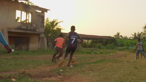 Ein-Junge-In-Einem-Orangefarbenen-Hemd-Dribbelt-Den-Ball-Durch-Andere-Spieler-Hindurch,-Mit-Heruntergekommenen-Gebäuden-Im-Hintergrund-Auf-Einem-Gemeinschaftsfußballplatz-In-Kumasi,-Ghana