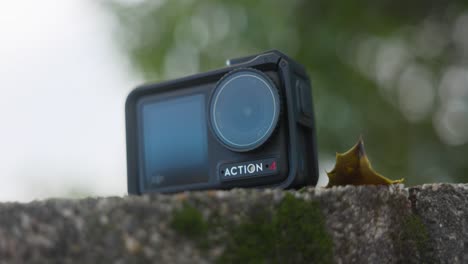 Toma-De-Producto-En-ángulo-Bajo-De-La-Cámara-Dji-Osmo-Action-4-Apoyada-En-Una-Pared-De-Piedra-Con-Fondo-De-árbol-Bokeh-Verde