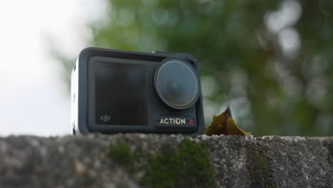 Low-Angle-Produktaufnahme-Der-DJI-Osmo-Action-4-Kamera,-Die-Auf-Einer-Steinmauer-Mit-Grünem-Bokeh-Baumhintergrund-Ruht