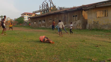 Boy-with-orange-shirt-falls-to-the-ground-as-others-carried-on-playing,-a-boy-with-yellow-shirt-dives-defending-the-imaginary-goal-made-of-two-rocks,-Kumasi,-Ghana