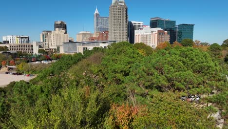 Aerial-reveal-of-Raleigh-skyline-from-behind-green-trees-during-autumn-day