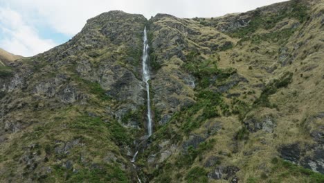 Scenic-aerial-view-panning-right-to-left-of-narrow,-free-flowing-waterfall-winding-through-rugged,-rocky-and-mountainous-landscape-in-the-remote-wilderness-of-New-Zealand-Aotearoa
