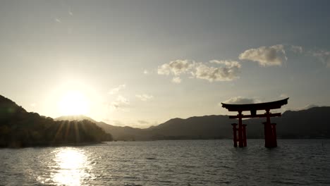 Silhouette-Des-Großen-Torii-Tors-Von-Itsukushima,-Das-Mit-Sonnenuntergangsfackeln-Im-Hintergrund-über-Der-Bergkette-Schwebt