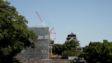 Der-Uto-Turm-Ist-Mit-Einem-Gerüst-Bedeckt,-Mit-Einem-Baukran-Für-Reparaturarbeiten,-Mit-Der-Burg-Kumamoto-Im-Hintergrund-Hinter-Bäumen-An-Einem-Klaren,-Sonnigen-Tag