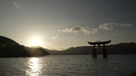 Malerische-Aussicht-Auf-Die-Silhouette-Des-Großen-Torii-Tors-Von-Itsukushima,-Das-Mit-Sonnenuntergangsfackeln-Im-Hintergrund-über-Der-Bergkette-Schwebt