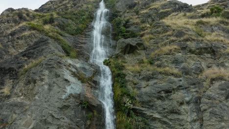 Close-up-aerial-view-of-picturesque-narrow-waterfall-falling-over-rugged,-rocky-landscape-in-the-remote-outdoor-wilderness-of-New-Zealand-Aotearoa