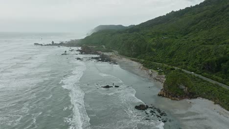 Reverse-aerial-ocean-view-of-wild-West-Coast-on-the-South-Island-of-New-Zealand-Aotearoa,-with-waves-rolling-into-shoreline-and-green,-tree-covered-landscape