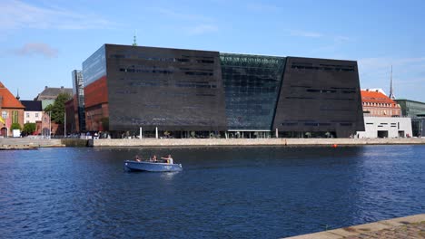 People-on-boat-navigating-canal-by-Royal-Danish-Library,-Copenhaguen,-Denmark